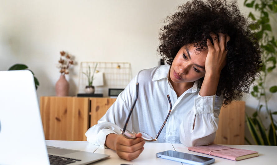 business woman holding her glasses away from her face and leaning her head in her hand, looking at laptop on desk