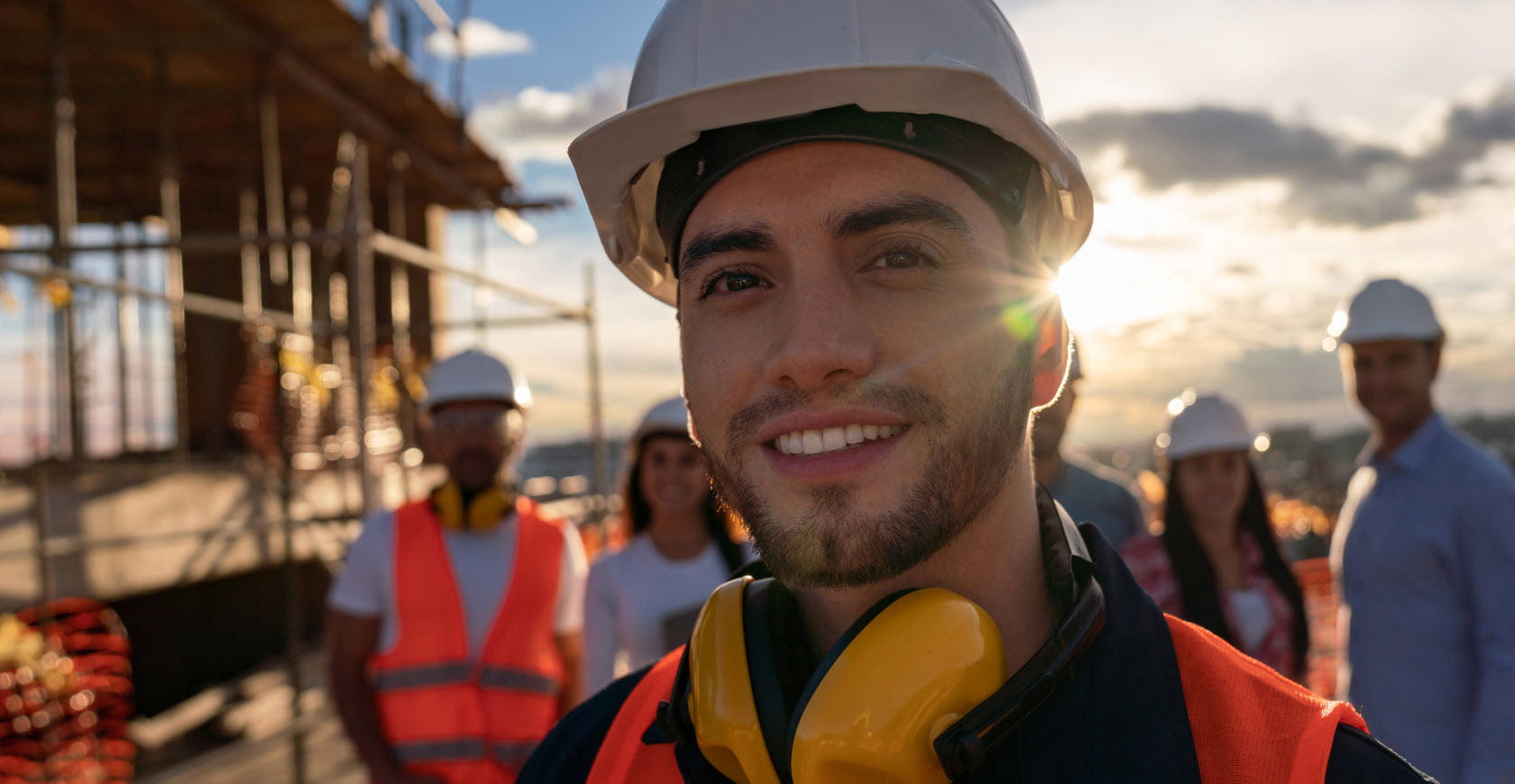 closeup of young Hispanic male construction worker wearing a safety helmet and smiling for camera. other construction workers are in the background.
