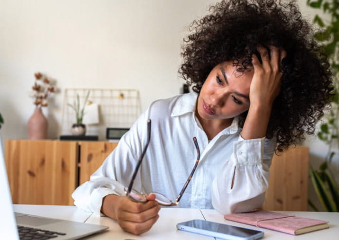 business woman holding her glasses away from her face and leaning her head in her hand, looking at laptop on desk