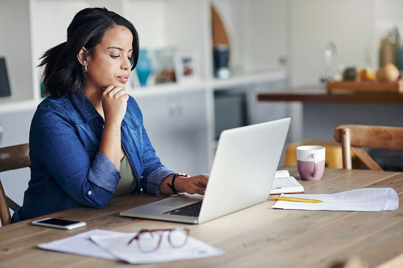 black woman probably in her thirties is sitting at dining room table looking at laptop